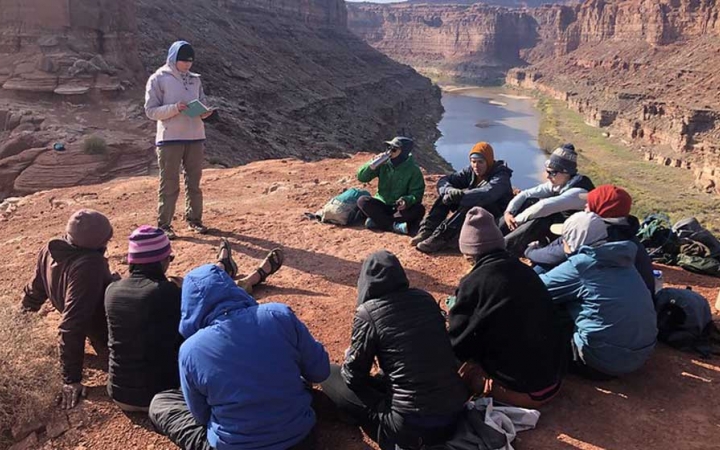 a group of students sit atop a rock high above a river listening to a reading from the outward bound readings book